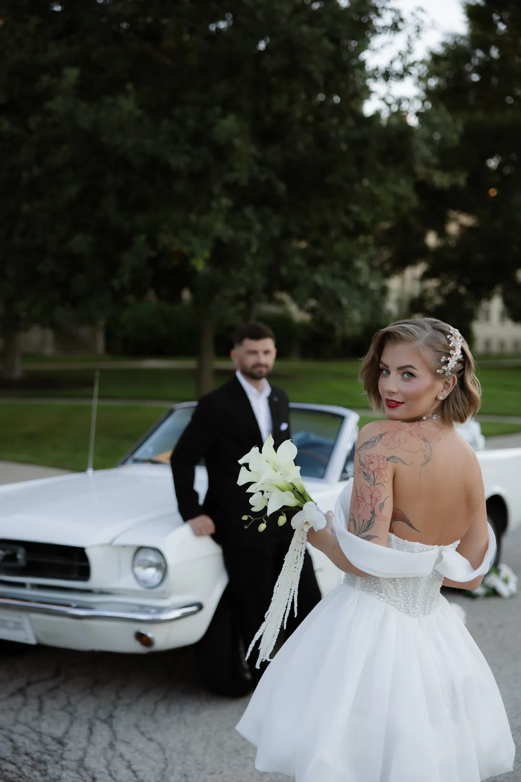 Bride glancing back over her shoulder, groom beside a vintage car